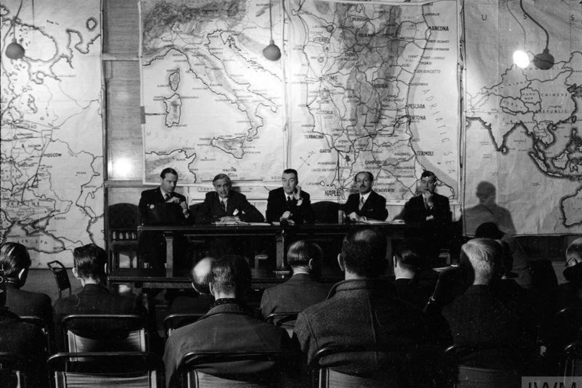 A historical black-and-white photograph of a panel of five men seated at a long table, addressing an audience, with large maps on the wall behind them.